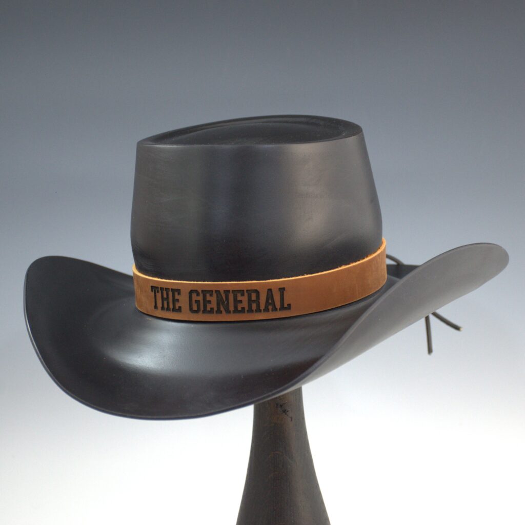 Black wooden cowboy hat with a smooth matte finish, displayed on a stand, featuring a tan leather band engraved with the words “THE GENERAL.”
