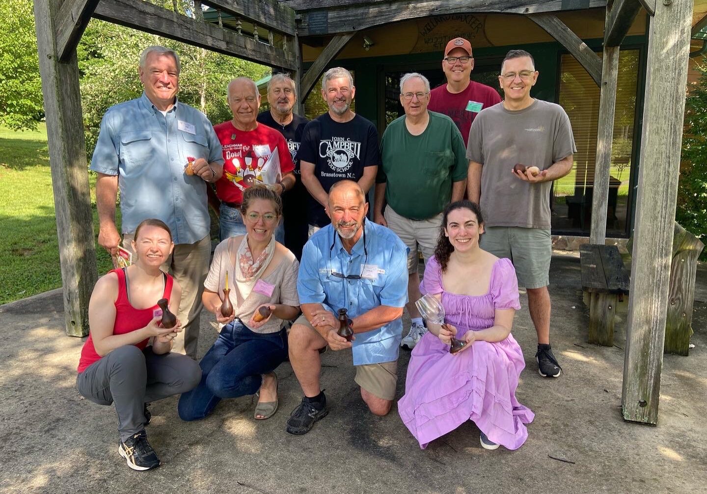 A group of woodturning students at John C. Campbell Folk School pose with their handcrafted wooden projects in front of the woodworking studio, smiling and celebrating their craftsmanship.