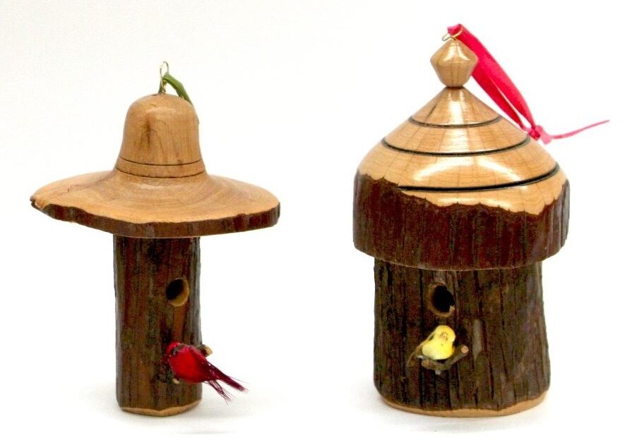 Two handcrafted wooden birdhouse ornaments with small decorative birds, each featuring unique designs and natural wood textures. One has a wide-brimmed hat roof, while the other has a tiered, polished top with a red ribbon for hanging.