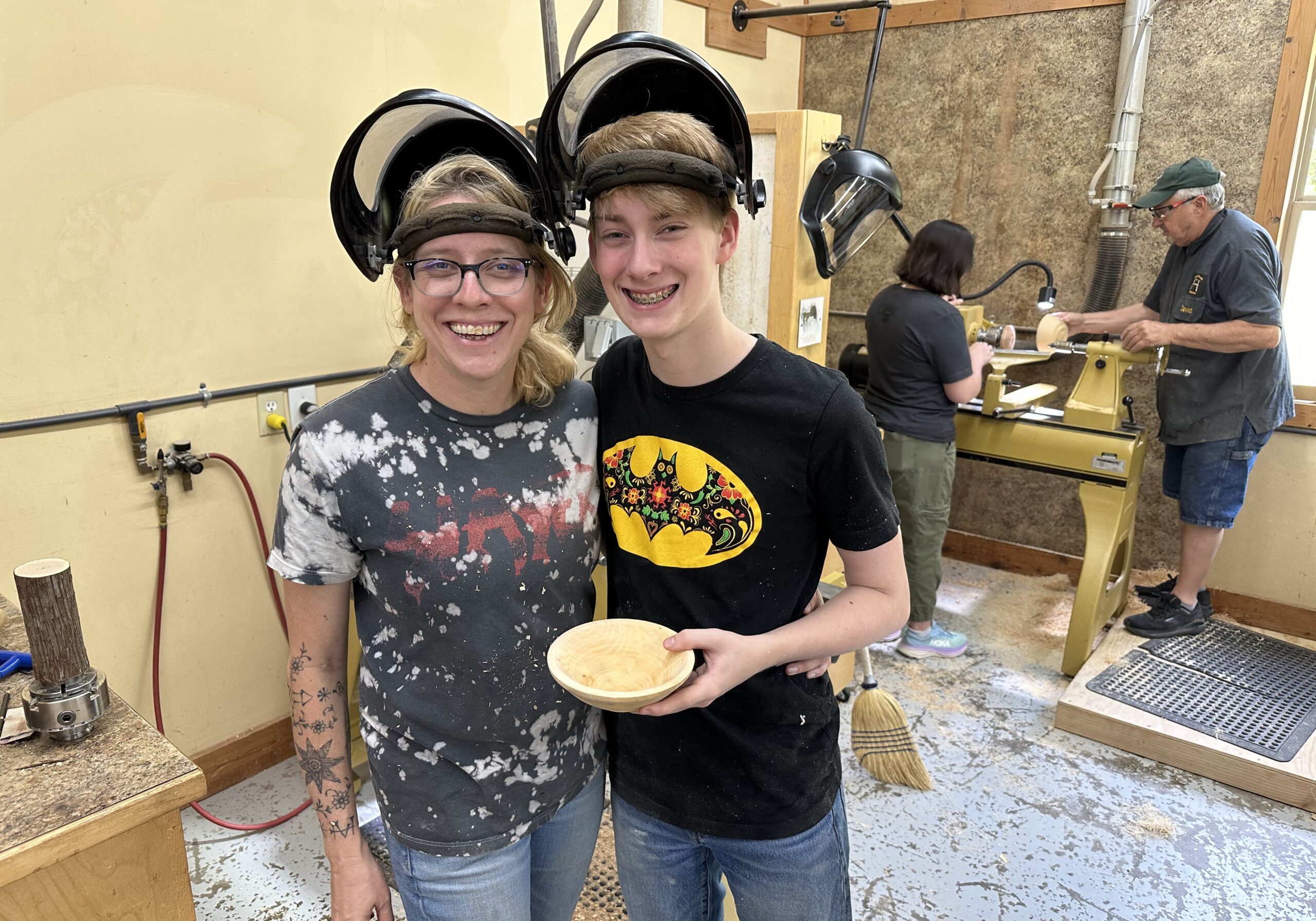 Two smiling students at John C. Campbell Folk School wear safety face shields while holding a handcrafted wooden bowl. In the background, other participants work on wood lathes in a sawdust-covered workshop.